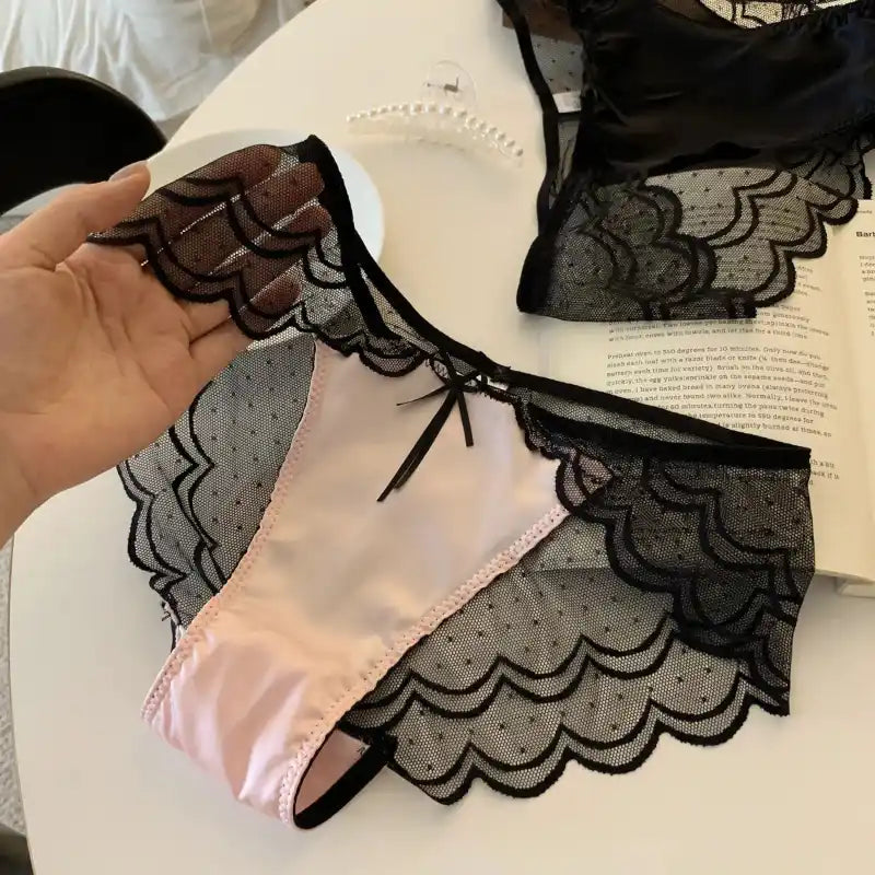 Hand holding blush pink bikini panties with black scalloped lace overlay and satin bow tie accent