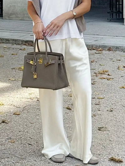 Woman outdoors holding a taupe Birkin-style bag, wearing ivory wide-leg satin pants with a white tee and gray sneakers