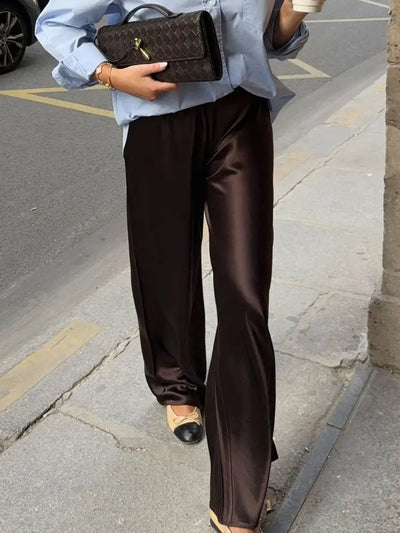 Woman walking in chocolate wide-leg satin pants paired with a light blue oversized shirt and two-tone ballet flats, holding a woven brown bag