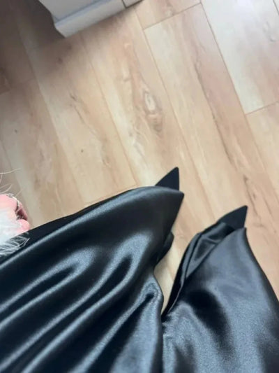 Close-up looking down at the flared hem of black wide-leg satin pants resting over pointed black flats on a wood floor