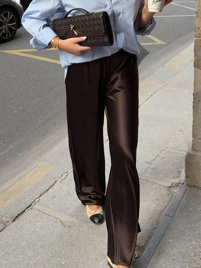 Full-length street view of chocolate wide-leg satin pants with a subtle ankle flare, worn with ballet flats and a blue shirt