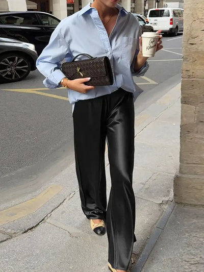 Woman on a city sidewalk wearing chocolate wide-leg satin pants, a blue button-down shirt, and two-tone flats, carrying a woven bag and a coffee cup