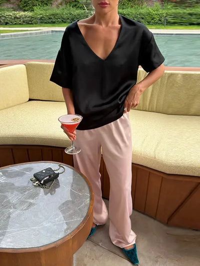 Woman by a poolside lounge area wearing blush wide-leg satin pants with a black V-neck top and teal flats, holding a cocktail glass