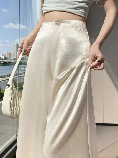 Woman holding ivory wide-leg satin pants at the hip showing drape and glossy finish outdoors