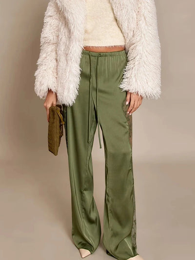 Front view of olive green satin wide-leg pants with lace panels, paired with a cream fuzzy coat and matching knit crop top, model holding an olive quilted bag