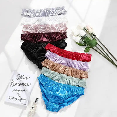 Flat lay of eleven satin panties in assorted colors beside white roses and perfume bottle