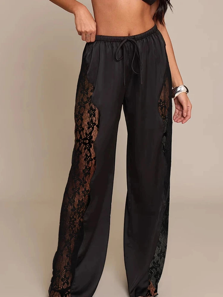 Black wide-leg satin pants with sheer floral lace side insets and elasticated drawstring waist, worn by a woman
