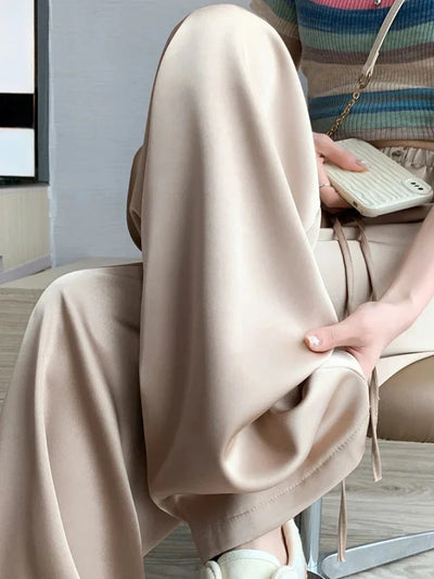 Side view of beige wide-leg satin pants draped over a seated figure showing full fluid silhouette and hem length