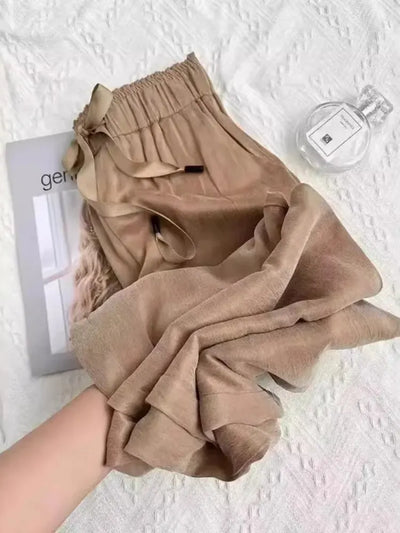 Folded camel wide-leg pants with bow-tie waistband displayed on white textured surface beside perfume bottle