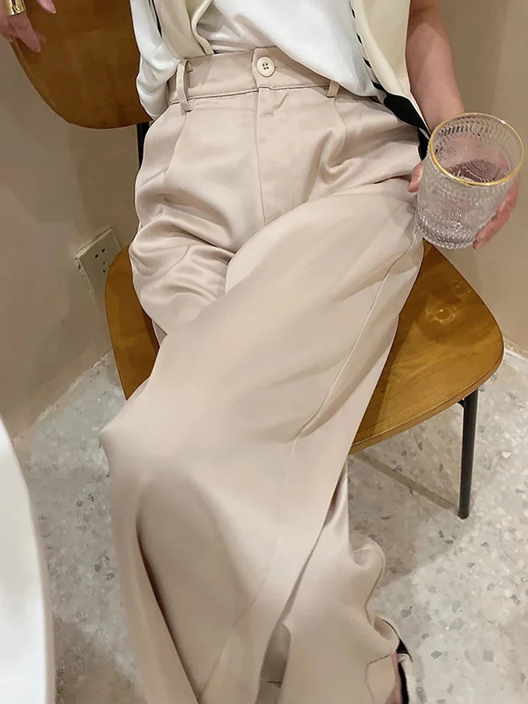 Woman seated on a wooden chair wearing high-rise beige wide-leg satin pants with a white top, holding a glass