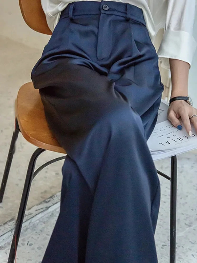Woman seated on wooden chair wearing high-rise navy pleated wide-leg satin pants with white blouse