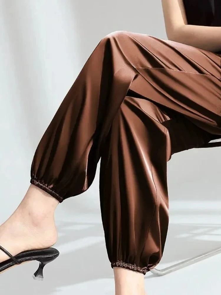 Woman seated wearing chocolate brown high-shine relaxed pants with elastic ankle cuffs and black heeled mules