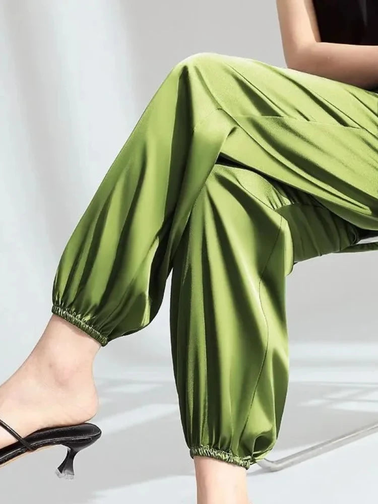 Woman seated wearing relaxed green satin pants with gathered elastic ankle cuffs and black heeled mules