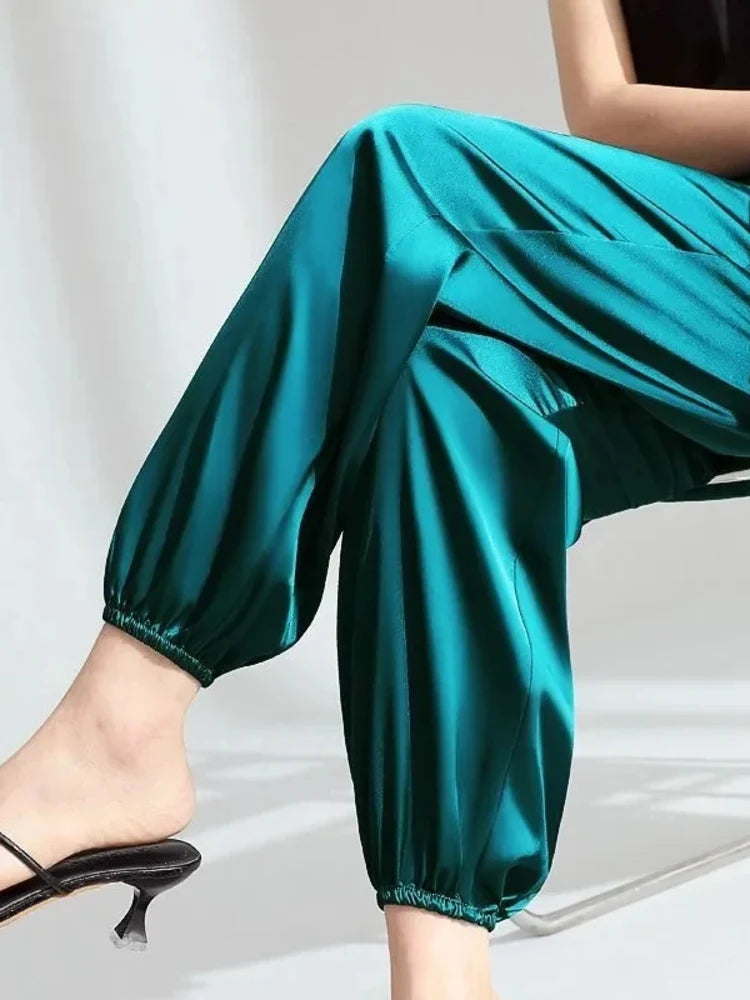 Woman seated wearing relaxed teal satin pants with elasticized ankle cuffs and black heeled mules