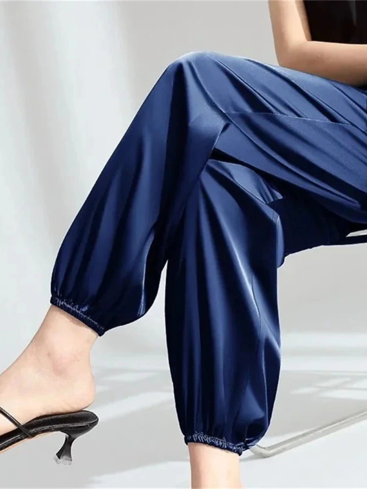 Navy blue satin gathered-ankle pants with elastic cuff worn with black strappy heels, seated pose showing fluid drape and tapered silhouette