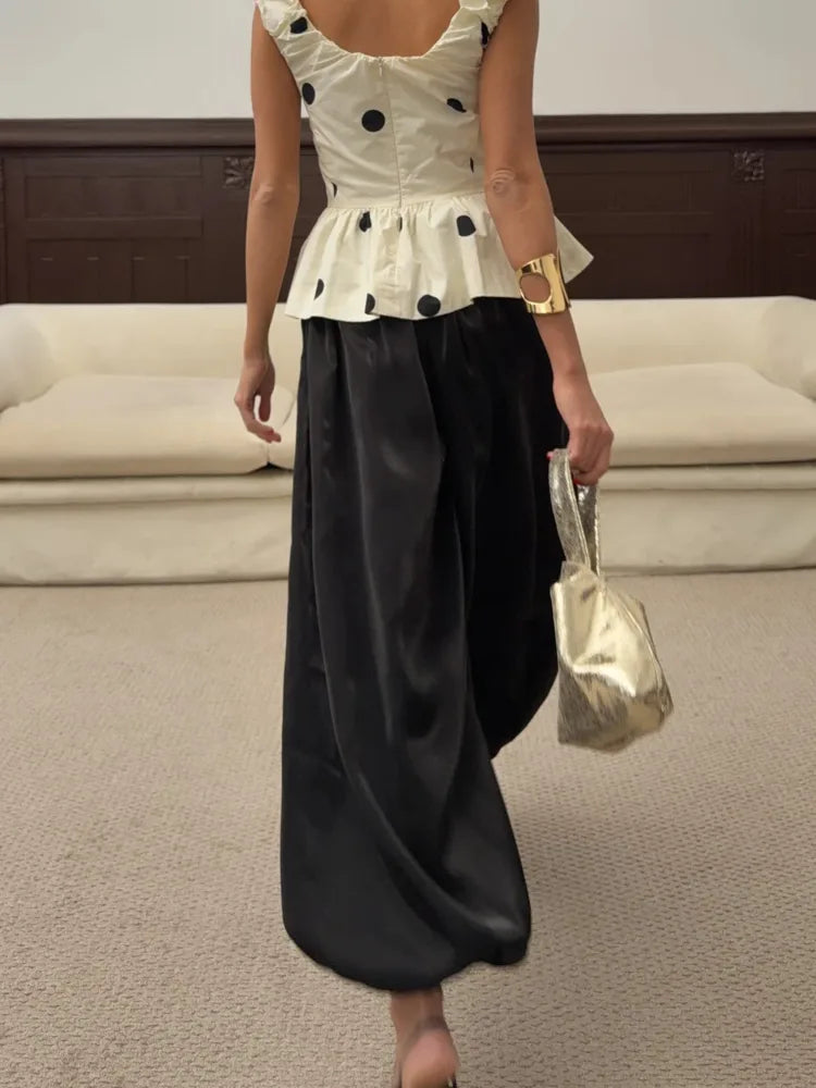 Back view of woman wearing black wide-leg satin pants with white polka-dot peplum top and gold clutch