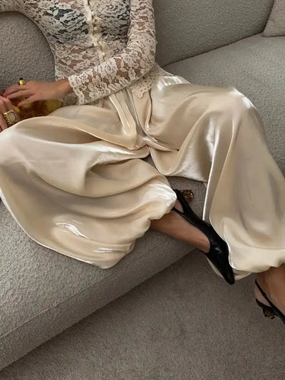 Woman seated on a sofa wearing flowing ivory satin wide-leg pants with a lace blouse and black heeled sandals
