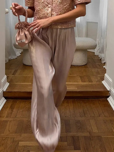 Woman in blush satin wide-leg pants styled with a pink embroidered top and a small drawstring bag