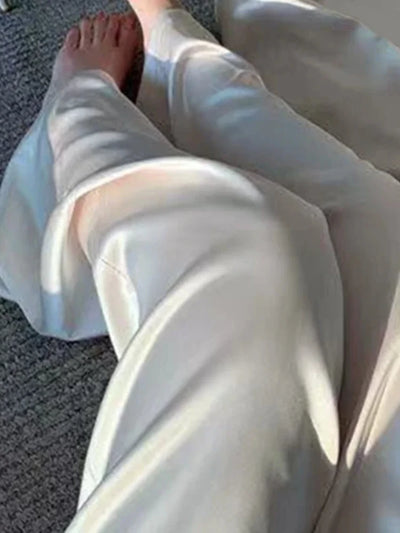 Ivory wide-leg satin pants with fluid drape seen from the side, wearer seated on floor barefoot