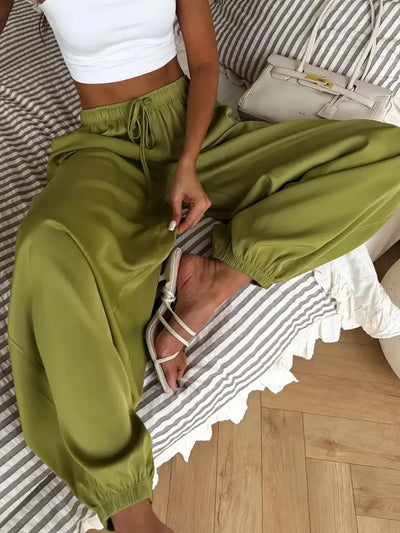 Close-up of olive green balloon satin pants with drawstring waist, worn seated with white strappy sandals