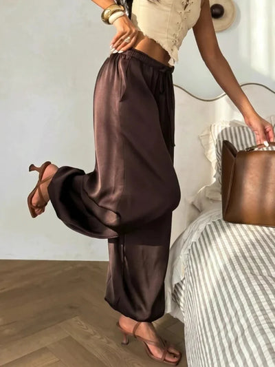 Close-up side view of dark chocolate fluid wide-leg pants with elasticated drawstring waist and glossy satin finish, styled with brown heeled sandals