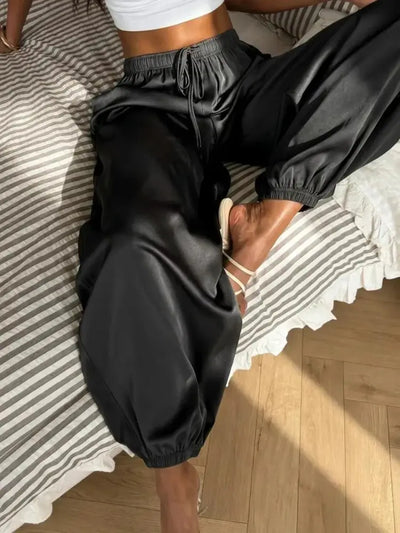 Woman lying on striped bedding wearing black satin jogger pants with strappy heeled sandals