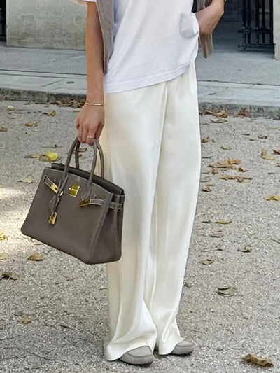 Woman holding a gray Hermes Birkin bag wearing off-white straight-leg satin pants and white tee