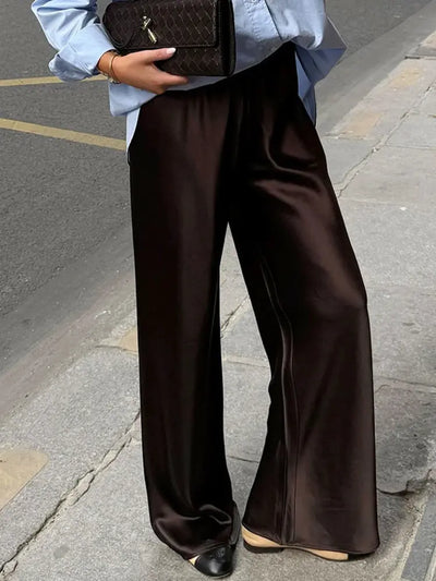Close-up of dark brown wide-leg satin pants with glossy finish and black-tipped ballet flats on a sidewalk
