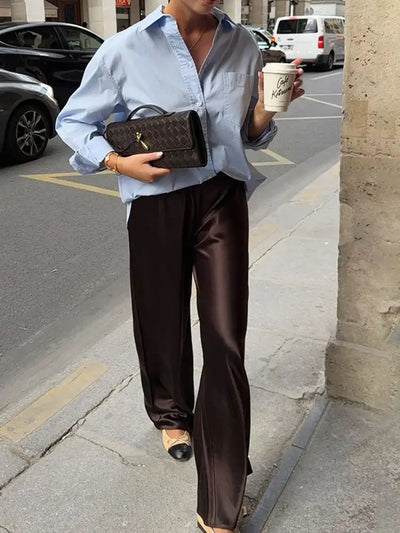 Woman walking in dark brown wide-leg satin pants and oversized blue shirt holding a coffee cup and woven clutch