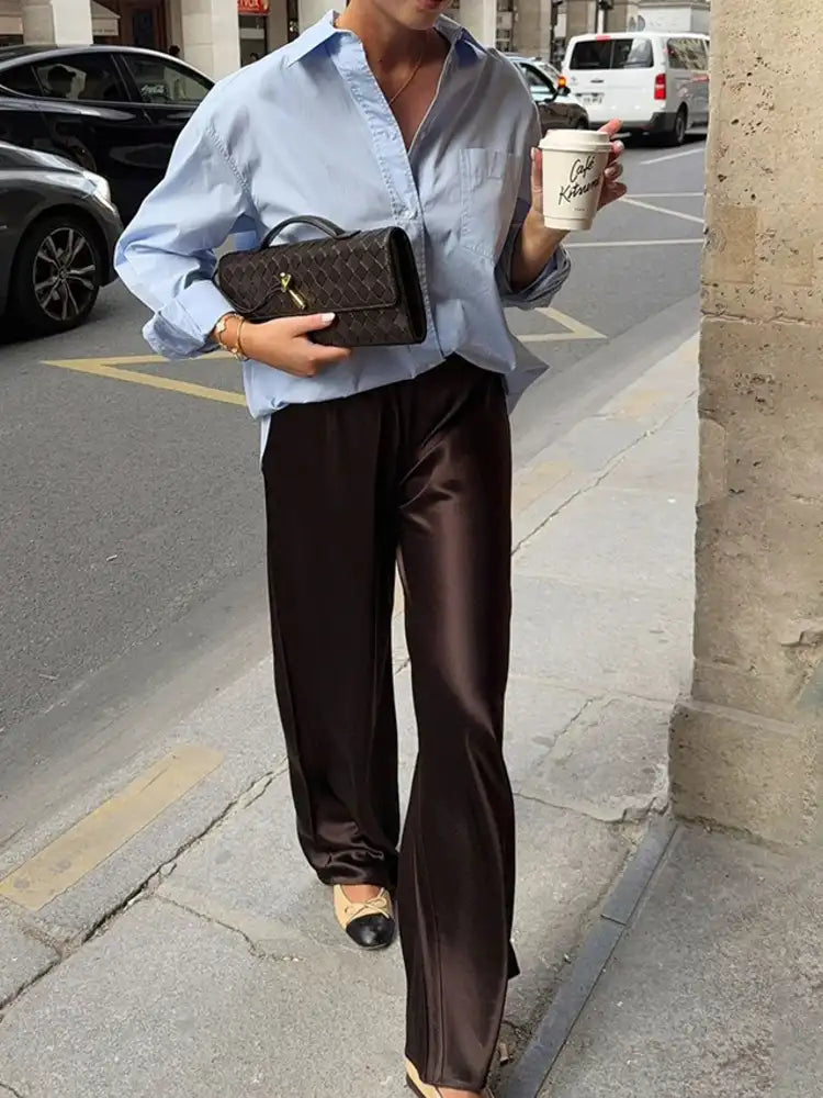Close-up of dark brown wide-leg satin pants with glossy finish and black-tipped ballet flats on a sidewalk