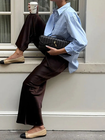 Woman leaning on a windowsill wearing dark brown wide-leg satin pants, blue shirt, and two-tone ballet flats