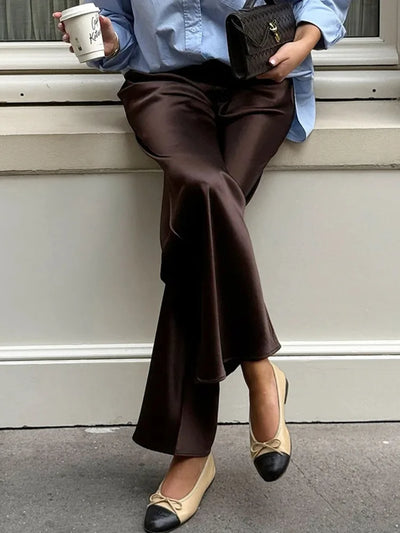 Close-up of the wide flared hem of dark brown satin pants with two-tone beige and black ballet flats