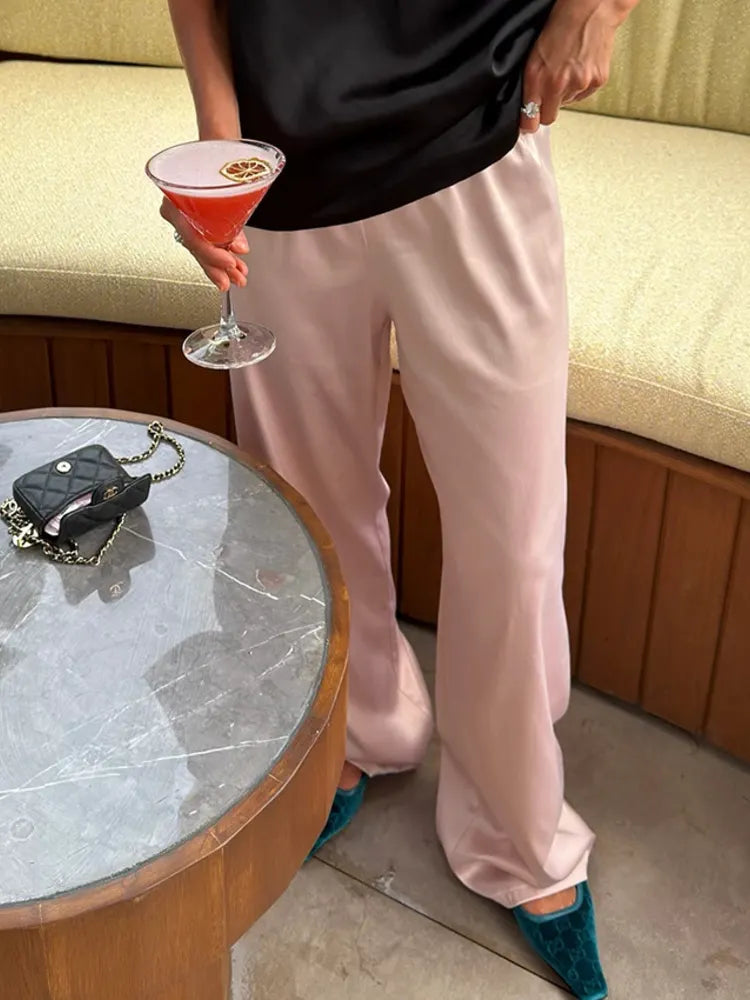 Person holding a pink cocktail wearing blush wide-leg satin pants with teal pointed-toe mules near a marble table