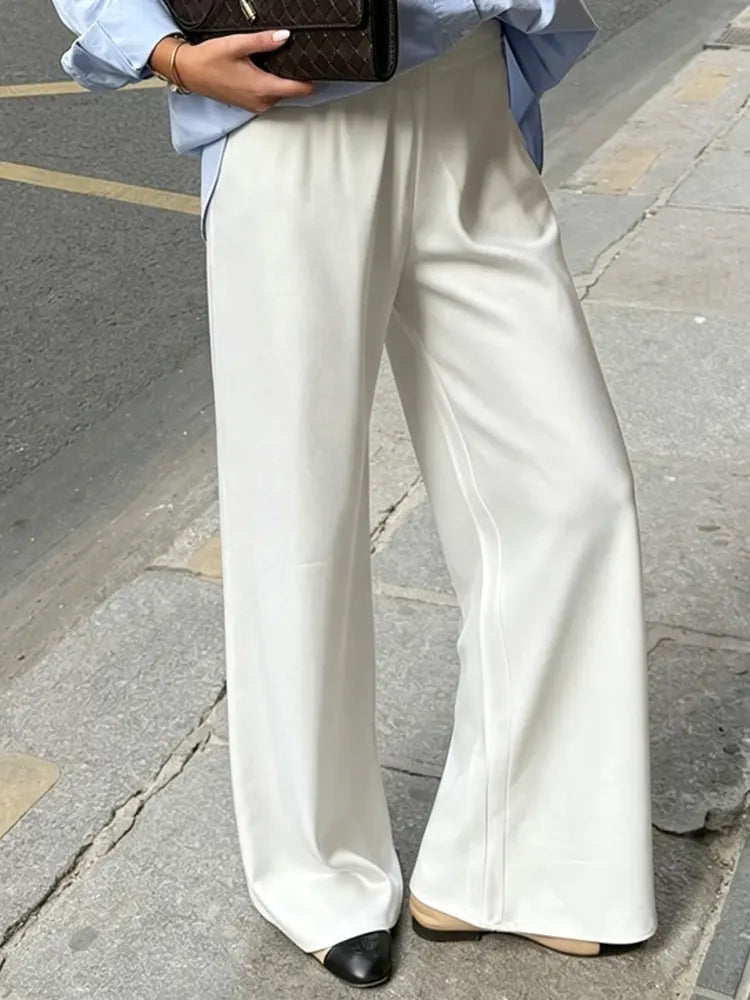 Woman wearing white straight-cut glossy wide-leg satin pants with a light blue oversized shirt and two-toned flats on a city sidewalk