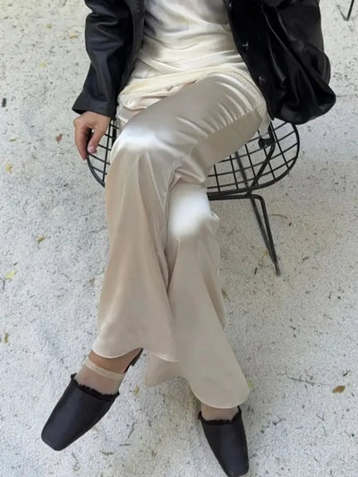 Seated woman in champagne satin straight-leg pants, black leather jacket and square-toe flats