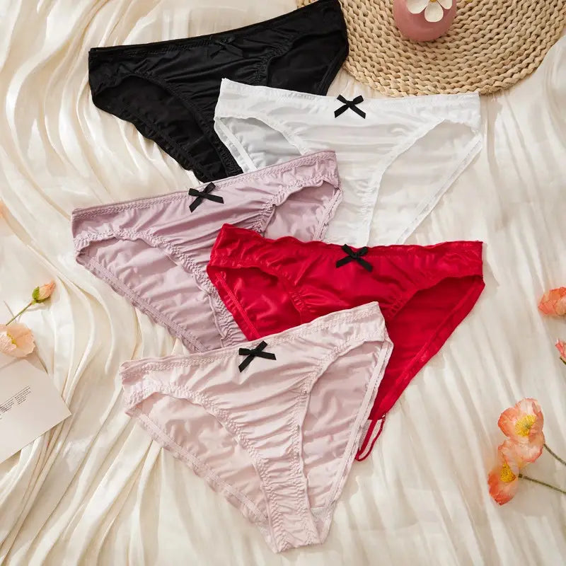 Five satin ruched bikini panties in black, white, lavender, red, and light pink laid flat on white satin fabric with small black bows