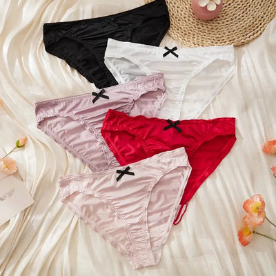 Five satin ruched bikini panties in black, white, lavender, red, and light pink laid flat on white satin fabric with small black bows
