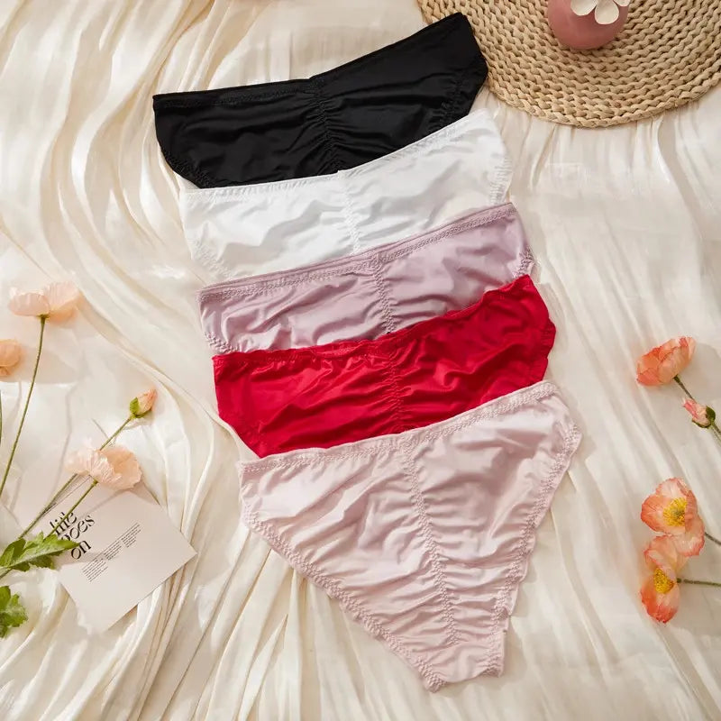 Five satin ruched bikini panties in black, white, lavender, red, and light pink laid flat on white satin fabric with small black bows