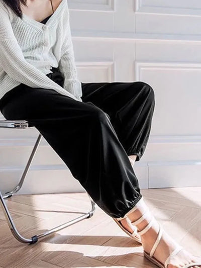 Woman seated on folding chair wearing black tapered satin pants with strappy sandals and white knit cardigan