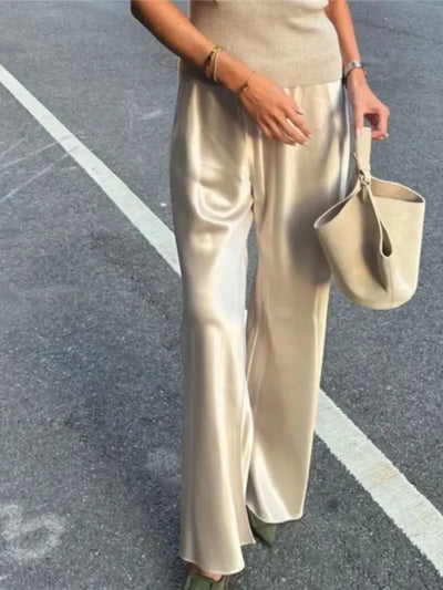 Woman wearing champagne wide-leg satin pants with high-shine finish, holding a beige bucket bag outdoors