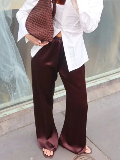 Person holding a woven brown bag wearing deep burgundy wide-leg satin pants and a white oversized shirt with open-toe slides