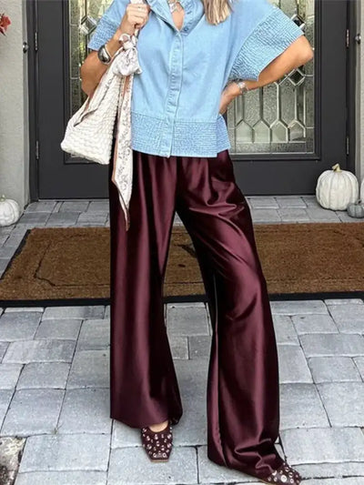 Woman in a blue denim top and deep burgundy wide-leg satin pants holding a cream woven bag outside a front door