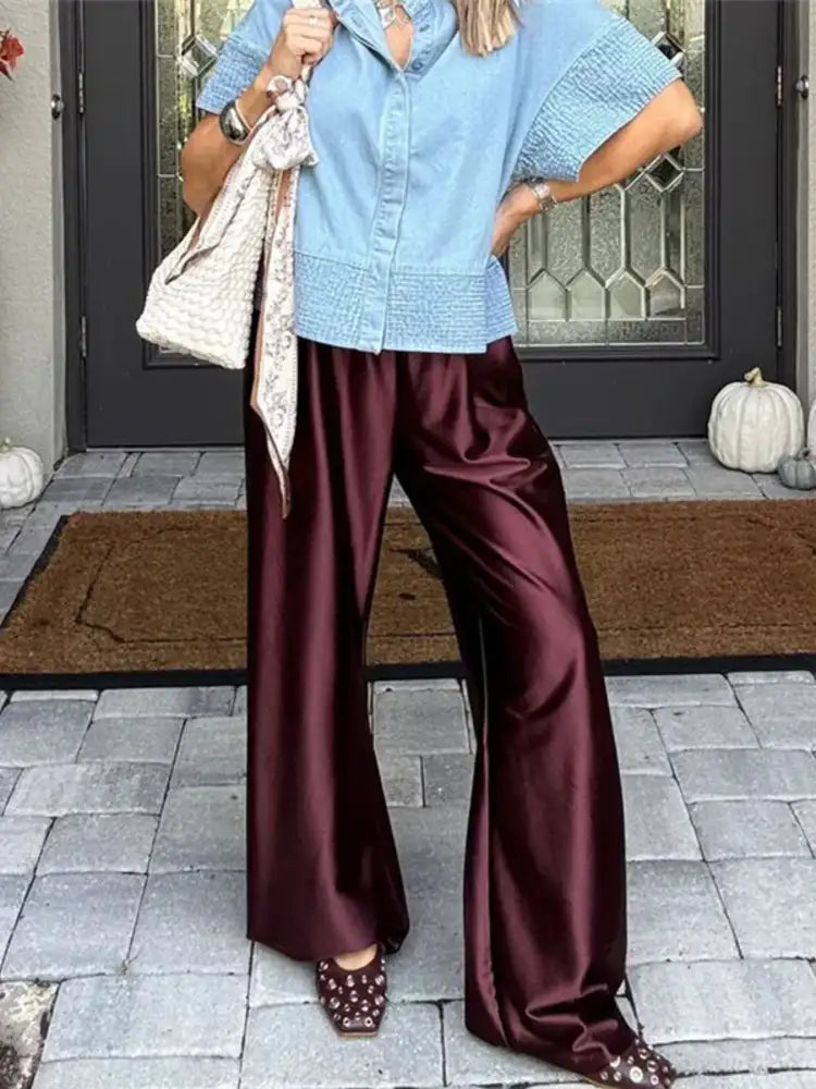 Person holding a woven brown bag wearing deep burgundy wide-leg satin pants and a white oversized shirt with open-toe slides