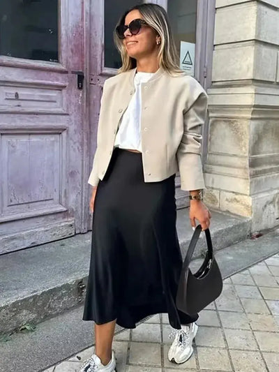 Woman wearing a black satin bias-cut midi skirt with a beige cropped jacket and white sneakers outdoors