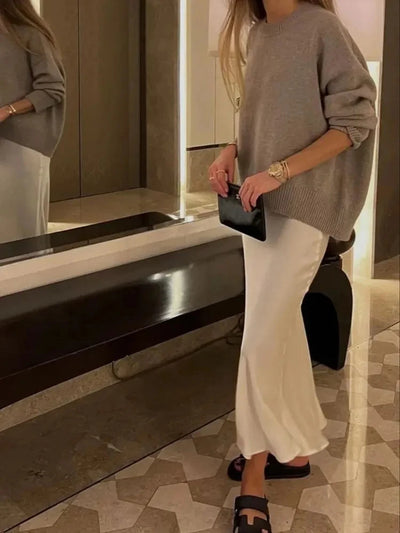 Woman holding a small black clutch wearing a white ivory satin midi skirt with a gray oversized knit sweater