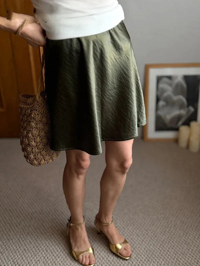 Side view of olive green flared mini satin skirt with gold sandals and straw bag