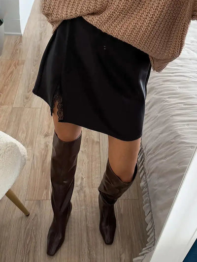 Overhead view of black satin mini skirt with lace split detail styled with dark brown knee-high boots and chunky knit sleeves
