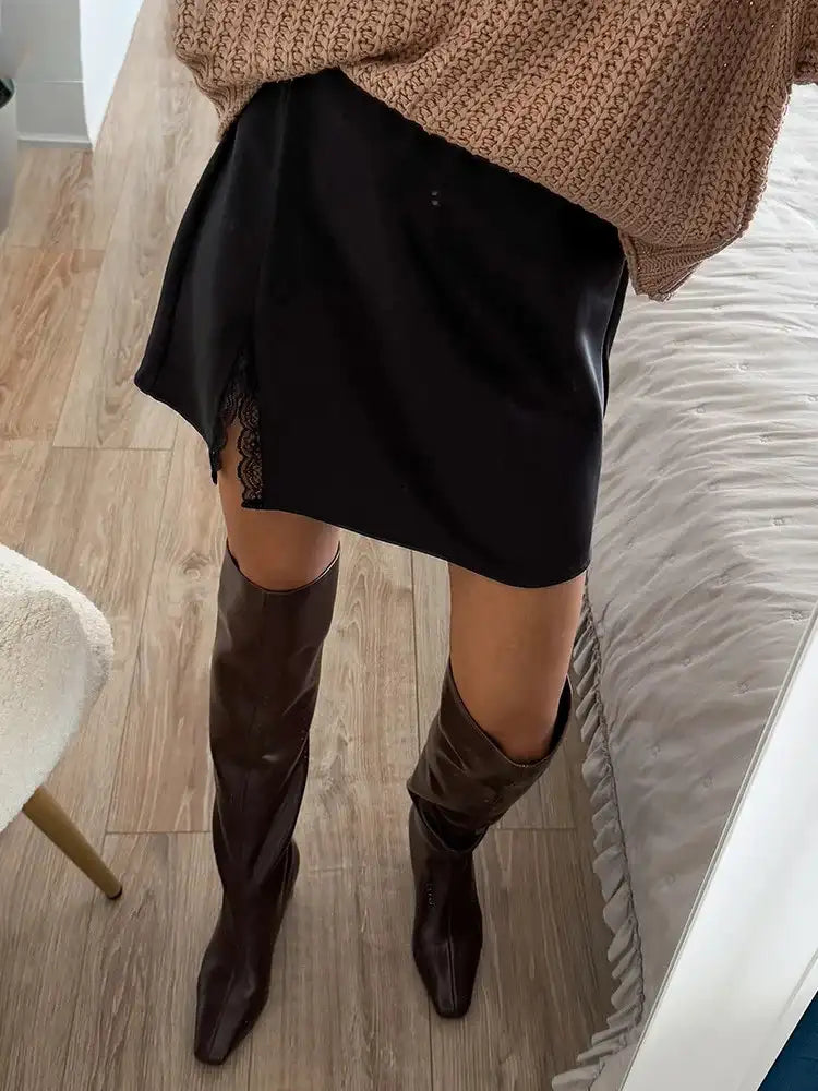 Close-up of black satin mini skirt with scalloped lace split hem worn with knee-high brown leather boots and camel knit sweater