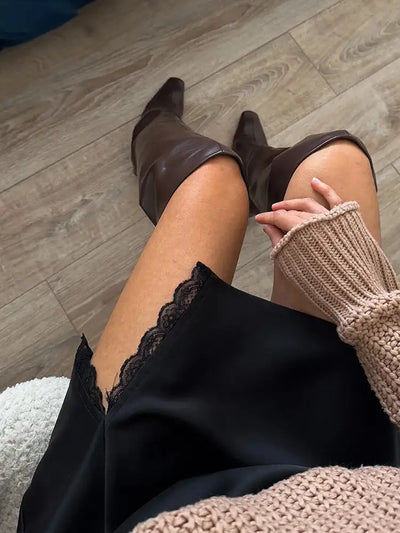 Seated close-up showing scalloped black lace trim on satin mini skirt split with brown leather knee-high boots visible