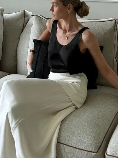 Woman seated on gray sofa wearing white maxi skirt and black satin tank top holding dark clutch